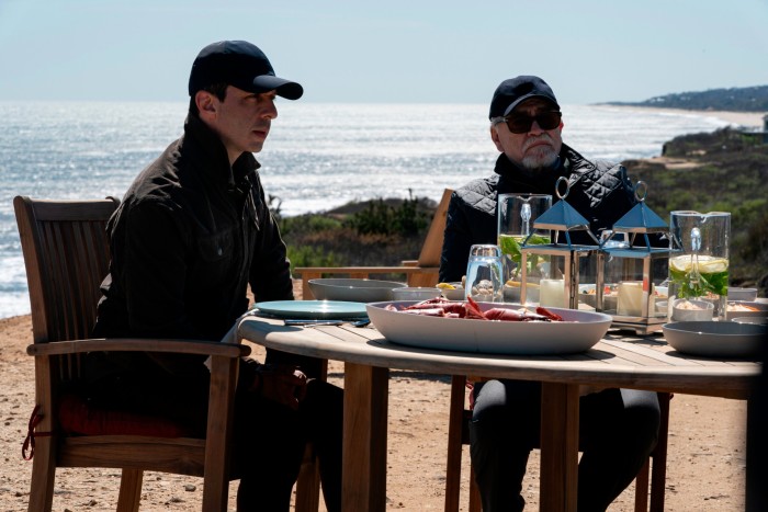 Two men sit at a table on a sunny beach. Both wear baseball caps