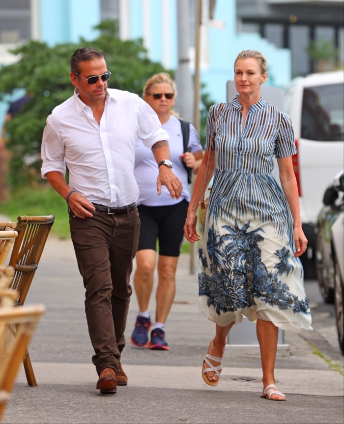 Lachlan and Sarah Murdoch walk on a street. He is in brown trousers and open-neck white shirt; she wears a patterned blue and white dress 