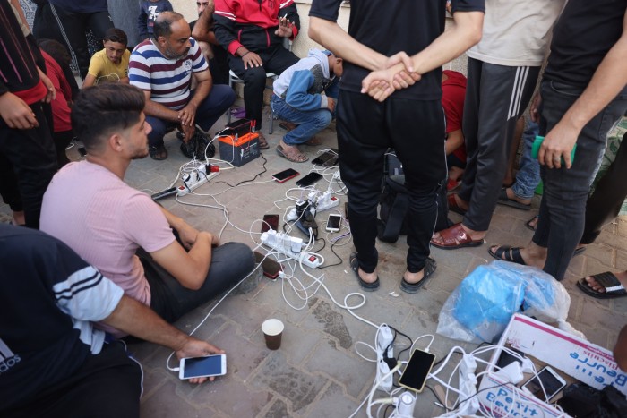 People sitting or standing on a pavement around a multi-socket extension cord from which they are charging their phones