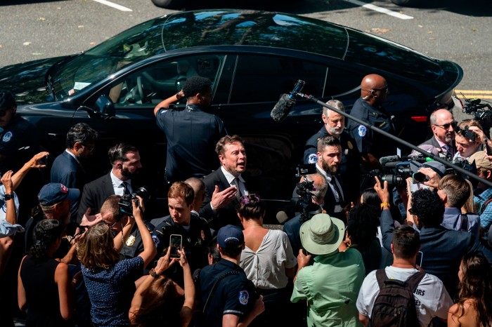 Man (Elon Musk) in a dark suit speaking in front of a black Tesla car as reporters and photographers surround him on the pavement