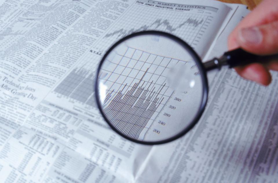 A person holding a magnifying glass above volume data and a stock chart printed in a financial newspaper.
