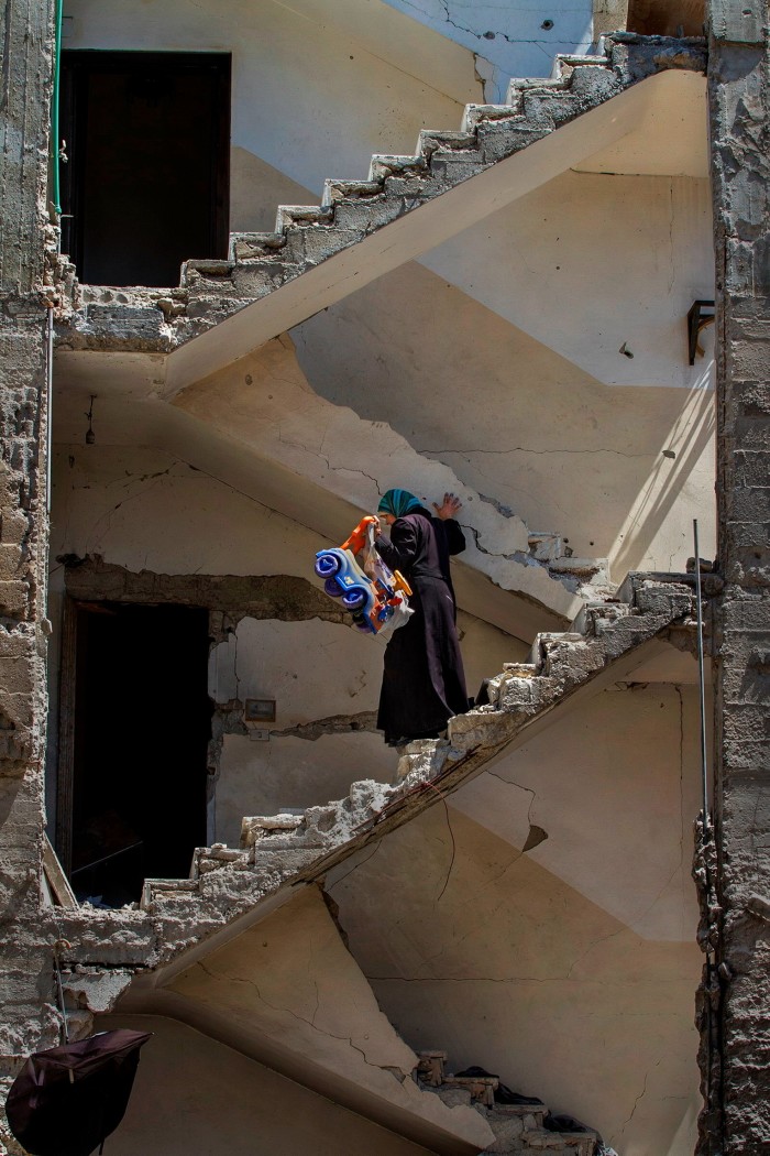 Woman in coat and headscarf descending staircase of home exposed after exterior wall has been bombed away. She is holding a child’s ride-on wheeled toy