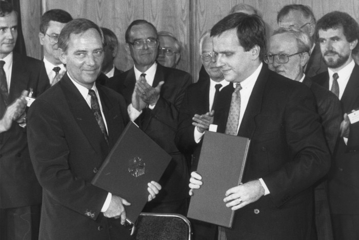 Two men in suits pose for cameras while each holding hard-backed document books